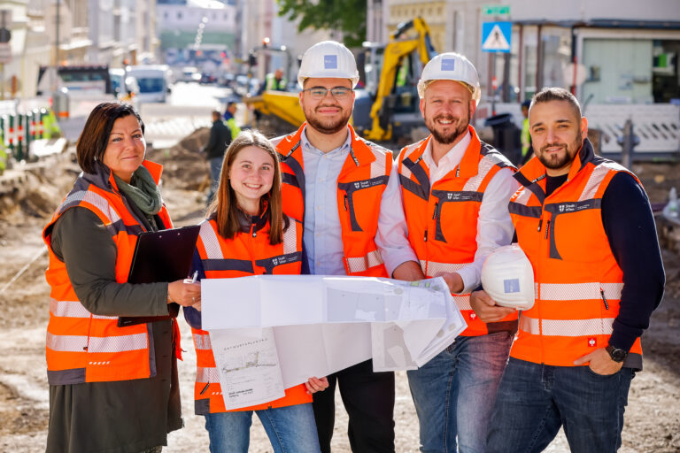 Eine Gruppe von Mitarbeitern und Mitarbeiterinnen der Stadt Wien steht lächelnd vor einer Baustelle. Sie halten gemeinsam einen großen Plan.