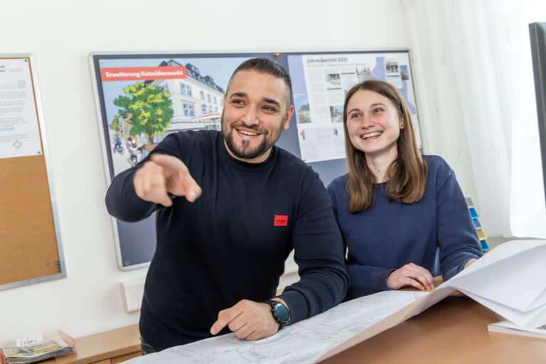 Ein Mitarbeiter und eine Mitarbeiterin der Stadt Wien schauen lächelnd nach Links, der Mitarbeiter zeigt in die Richtung