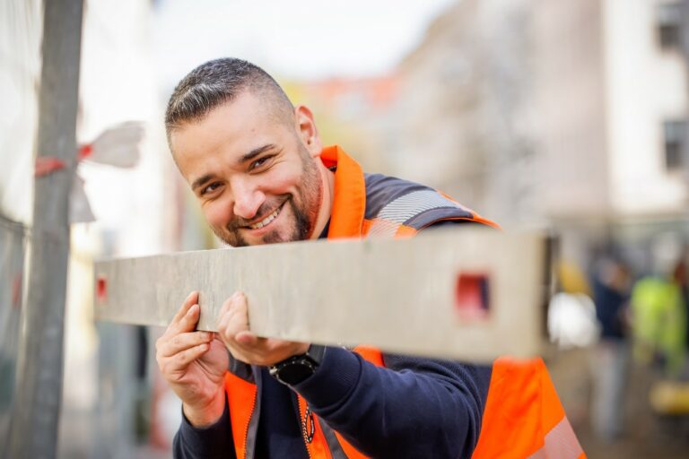 Werkmeister Aleksandar Begovic hält mit scherzhaftem Lächeln eine Abziehlatte aus Aluminium in beiden Händen, als würde er auf etwas in der Entfernung zielen.